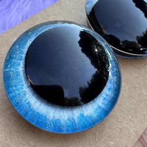 May include: Two round, glossy black and blue decorative glass pieces. The outer ring is a vibrant blue with a shimmering, speckled effect. The center is a deep, reflective black. The pieces are displayed on a light brown surface.