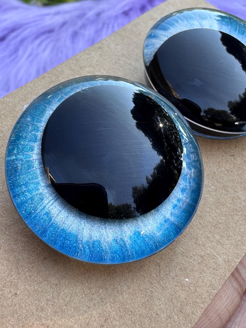 May include: Two round, glossy black and blue decorative glass pieces. The outer ring is a vibrant blue with a shimmering, speckled effect. The center is a deep, reflective black. The pieces are displayed on a light brown surface.