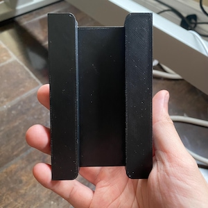 May include: A black phone holder with a U-shaped design. The holder is made of a matte material and features two vertical side supports and a central back panel. The holder is designed to securely hold a phone.