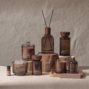 Op de afbeelding: Een collectie van huisparfumartikelen van donkerbruin glas, waaronder een geurstokjesdiffuser, diverse kaarsen met deksel, een klein flesje etherische olie en een rechthoekige doos, allemaal voorzien van het label Blossom of Freedom.