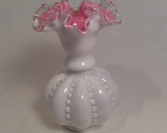 May include: A white glass vase with a ruffled pink and clear rim. The vase has a bulbous body with vertical beaded accents and a rounded base. The top of the vase flares out, creating a decorative and elegant design.