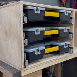 May include: A wooden cabinet with multiple pull-out drawers. The drawers are black with gray lids and yellow handles. The cabinet is made of light-colored wood and is designed for storage.