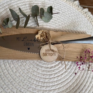 Könnte beinhalten: Ein Tortenheber mit gezahntem Rand und Holzgriff, graviert mit den Worten "Mit Liebe Gebacken" und einem Herzsymbol. Ein Holzanhänger mit "Happy Birthday" ist befestigt. Der Heber liegt auf einem Holzbrett, verziert mit Trockenblumen und Grün.