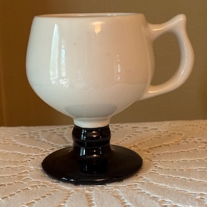 May include: A white ceramic goblet-shaped mug with a black base and stem. The mug has a rounded bowl and a curved handle. The mug is sitting on a white lace tablecloth. The mug is likely used for drinking.