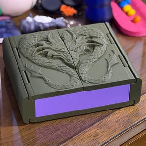 May include: An olive green hinged box with a purple interior. The top of the box features a detailed relief of two intertwined dragons. The box is rectangular and appears to be made of plastic or resin. The box is sitting on a wooden surface.