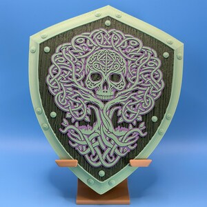 May include: A decorative shield featuring a skull design within a tree-like pattern. The shield has a green border and a dark wood-like center, with purple and green accents. It is mounted on a brown stand against a blue background.