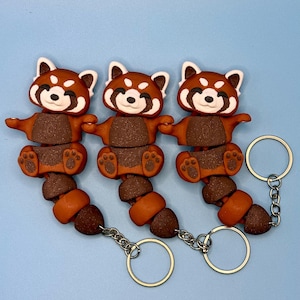 May include: Three red panda keychains. Each keychain features a brown and orange red panda with a white face and a silver keyring. The keychains are displayed against a light blue background.