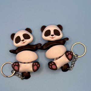 May include: Two panda bear keychains. Each keychain features a white panda with black accents, including ears, arms, and legs. The pandas are holding hands and have pink paw pads. Each keychain has a silver chain and ring.