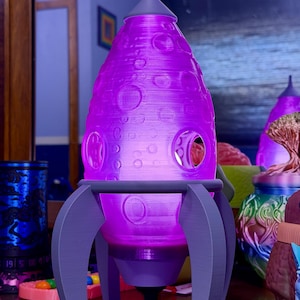 May include: A purple rocket-shaped lamp with a textured surface and circular cutouts. The lamp has gray legs and a pointed top. The lamp is lit from within, casting a purple glow. The lamp is a decorative item.