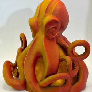 May include: A 3D-printed octopus sculpture in shades of orange and yellow. The tentacles curl around the body, and the eye sockets are detailed. The sculpture is likely made of plastic and sits on a white surface.