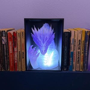 May include: A fantasy-themed bookshelf arrangement featuring a glowing dragon sculpture in a black frame, flanked by purple dragon wing bookends. Books of various sizes and colors are between the dragon and the bookends.