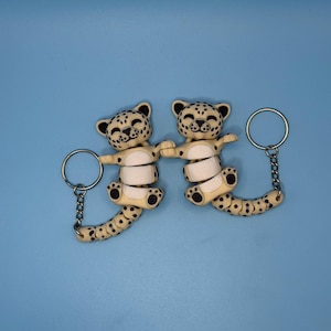 Peut inclure: Deux porte-clés représentant des léopards souriants avec des taches noires sur un corps crème. Les porte-clés ont des chaînes et des anneaux de couleur argentée. Le fond est bleu clair uni.