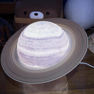 May include: A Saturn-shaped lamp with a white and gray planet and a light brown ring. The lamp is on a wooden surface, with a white cord. A brown bear-shaped night light and a white sphere are in the background.