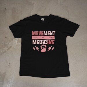 Pode incluir: T-shirt preto com a frase rosa "MOVEMENT IS MEDICINE". O design inclui um kettlebell e raios. A camisa &eacute; feita de um material macio, ideal para os entusiastas do fitness.
