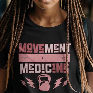 Pode incluir: Camiseta preta com o texto "MOVEMENT IS MEDICINE" em letras rosa e brancas. Um haltere rosa e um raio gr&aacute;fico est&atilde;o abaixo do texto. A camiseta tem gola redonda.