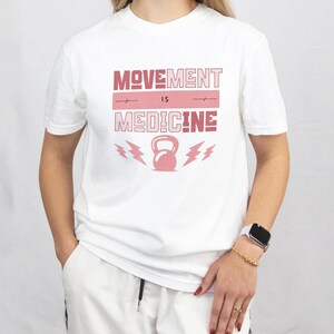 Pode incluir: Camiseta branca com a frase "MOVEMENT IS MEDICINE" em rosa. O design inclui um kettlebell rosa e raios. A camiseta &eacute; de gola redonda.
