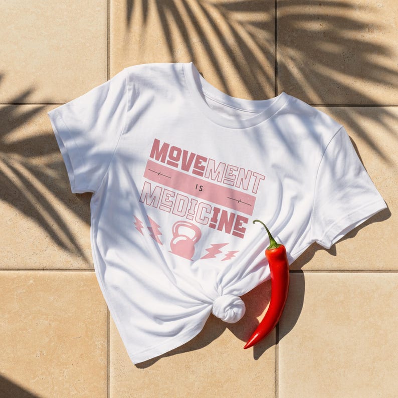 Pode incluir: Camiseta branca com a frase "MOVEMENT IS MEDICINE" em rosa. O design inclui um kettlebell e raios. Uma pimenta vermelha repousa sobre a superf&iacute;cie de azulejos ao lado da camiseta.
