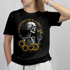 May include: Black t-shirt featuring a detailed graphic of a skeleton holding a butterfly, surrounded by sunflowers. The text "LIFE IS BETTER WHEN YOU'RE ALIGNED" is arched around the top. The design is in shades of yellow, white, and black.
