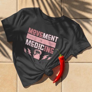 Pode incluir: Camiseta preta com a frase "MOVEMENT IS MEDICINE" em rosa e branco. O design inclui um kettlebell e gr&aacute;ficos de raios. Um piment&atilde;o vermelho repousa sobre a superf&iacute;cie de azulejos ao lado da camiseta.