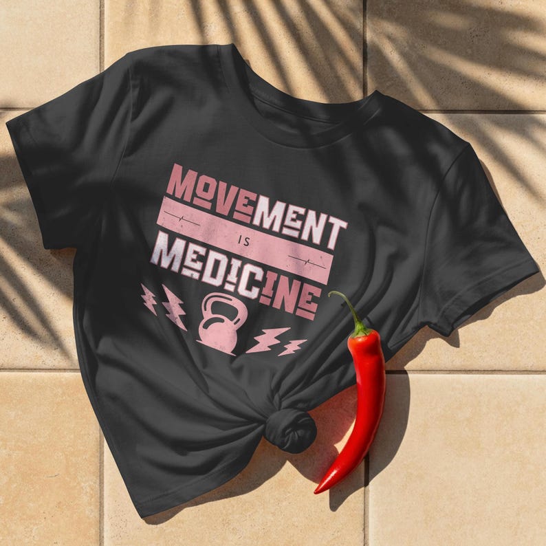 Pode incluir: Camiseta preta com a frase "MOVEMENT IS MEDICINE" em rosa e branco. O design inclui um kettlebell e gr&aacute;ficos de raios. Um piment&atilde;o vermelho repousa sobre a superf&iacute;cie de azulejos ao lado da camiseta.
