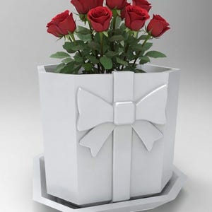 May include: A white hexagonal planter shaped like a gift box with a raised bow and ribbon design on the front. The planter sits on a matching hexagonal saucer and contains a bouquet of deep red roses with green leaves extending from the top.