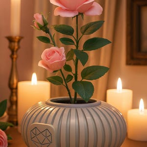 May include: A decorative arrangement featuring a pink rose plant in a striped vase. The vase has a geometric design on the side. Lit candles and a candle holder are in the background, creating a warm ambiance.
