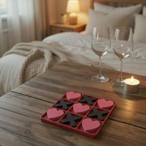 May include: A wooden table with a red tic-tac-toe game featuring pink hearts and black X's. Two wine glasses and a lit candle are on the table. A bed with white linens is in the background.