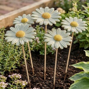 May include: Five decorative garden stakes featuring sculpted daisy flowers with pale green petals and textured yellow centers. Each flower is attached to a thin, straight wooden stem, designed to be inserted into the soil for outdoor display.