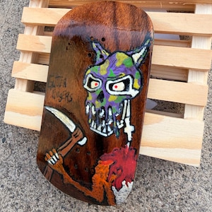 May include: A wooden skateboard deck with a skull and scythe design. The skull is painted in purple, green, and white, with red eyes. The scythe is held by a brown arm. The text "Gutter Knifers" is visible on the deck.