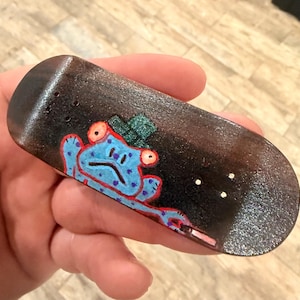 May include: A miniature finger skateboard with a black surface and a cartoon frog design. The frog is blue with red accents and a green hat. The skateboard has three white dots and is held in a hand.