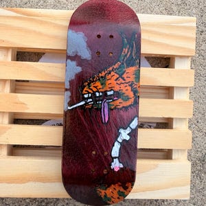 May include: A burgundy fingerboard with a colorful graphic design. The artwork features an orange creature with a long tongue, a white bone-like structure, and gray clouds. The fingerboard is placed on a wooden pallet.