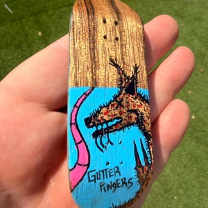 May include: A miniature wooden skateboard deck with a natural wood grain top. The bottom features a blue background with a cartoon wolf illustration and the words "Gutter Fingers" in white. A pink tail extends from the wolf.