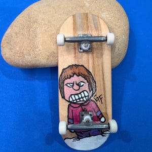 Può includere: Uno skateboard in legno con un'illustrazione dipinta a mano di un personaggio dei cartoni animati arrabbiato con una camicia rossa. Lo skateboard ha truck argentati e ruote bianche. Una grande pietra liscia e marrone chiaro poggia dietro lo skateboard su una superficie blu.