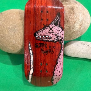 May include: A wooden fingerboard with a red stain and a pink and black illustration of a wolf-like creature. The text "GUTER XX FINGERS" is written in black. The fingerboard is a miniature skateboard.