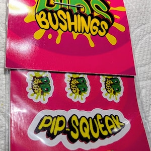 May include: A pink package with the words "Dibs Bushings" in green and yellow. Below are three stickers with a "B" design and the words "Pip-Squeak". A small orange and green object is at the bottom.