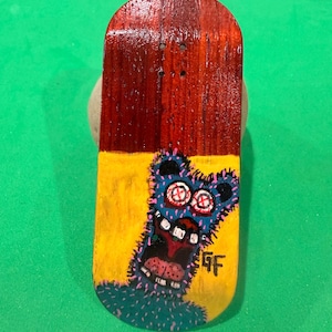 May include: A small, wooden fingerboard with a painted design. The top portion is stained a dark reddish-brown, while the bottom half is yellow with a blue monster illustration. The monster has large, red-and-white eyes and a wide, open mouth.