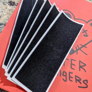 Puede incluir: Una pila de tiras de cierre de gancho y bucle negras con bordes blancos, dispuestas sobre una superficie roja. Las tiras son rectangulares y tienen una superficie texturizada. La superficie roja tiene texto y un gráfico.