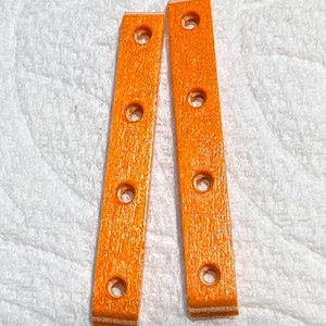 May include: Two orange wooden strips with six evenly spaced holes. The strips have a textured surface and are set against a white background. The color is a vibrant, solid orange.