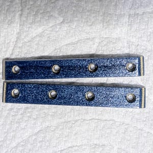 May include: Two rectangular, blue wooden pieces with six evenly spaced holes. The wood has a textured appearance and a metallic sheen. The edges are trimmed with a thin, light-colored border. The pieces are arranged parallel to each other.