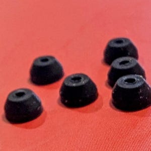 May include: A collection of seven black earbud tips, each with a conical shape and a central opening. The tips are arranged on a red surface, providing a contrasting background. These are likely replacement parts for audio devices.