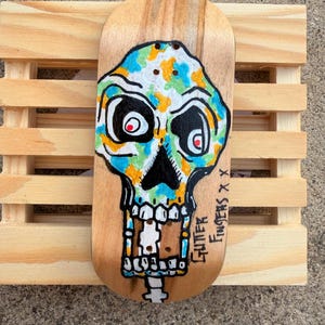 May include: A wooden fingerboard featuring a hand-painted skull design. The skull is painted with blue, green, and orange accents, with black outlines and red eyes. The text "Glitter Fingers X X" is written on the side. The fingerboard rests on a small wooden pallet.