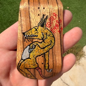 May include: A wooden fingerboard with a yellow monster illustration. The monster has a long snout, a silver belly, and a red tail. The words "GUTER FINGER" are written in red.