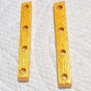 May include: Two rectangular wooden pieces with a golden finish. Each piece has four evenly spaced holes. The wood grain is visible, adding texture. The pieces are likely components for a craft or DIY project.