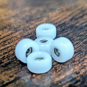 May include: A cluster of five small, light blue skateboard wheels arranged on a wooden surface. Each wheel has a rounded shape with a central metal bearing visible. The wheels are clean and appear unused.