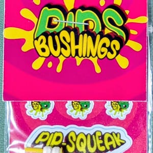 Puede incluir: Un paquete de bujes de monopatín. La tarjeta superior tiene el texto "Dibs Bushings" en verde y amarillo sobre un fondo rosa. La tarjeta inferior tiene el texto "Pip-Squeak" y pequeñas pegatinas.