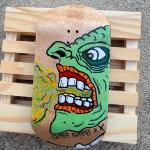 May include: A wooden skateboard deck featuring a green, cartoonish monster face with an open mouth and white teeth. The monster is painted on a light brown background. The words "GUTTER FINGERS XX" are written in black at the bottom.