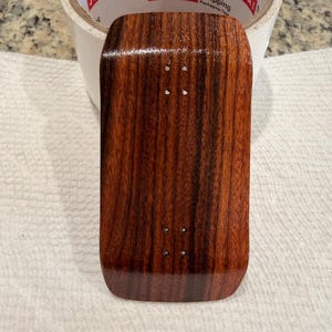 May include: A small, rectangular wooden fingerboard deck with a rich, dark brown grain pattern. The deck has four small holes and a glossy finish. The rounded edges and smooth surface suggest a high-quality, handcrafted item.