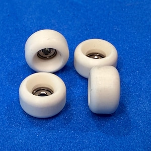 May include: Four small, white roller skate wheels with a silver bearing visible in the center of each. The wheels are arranged on a blue surface, showcasing their rounded shape and design.
