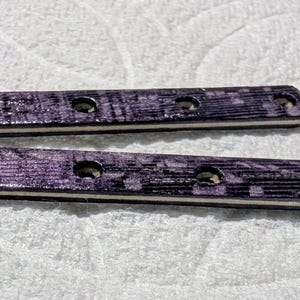 May include: Two rectangular wooden pieces with a purple and black grain pattern. Each piece has four evenly spaced holes. The edges are a light tan color. The pieces are likely decorative or functional elements.
