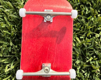 Könnte beinhalten: Ein rotes Fingerboard-Skateboard mit silbernen Trucks und weißen Rollen. Das Miniatur-Skateboard wird von oben gezeigt und liegt auf einem Bett aus grünem Kunstrasen. Das Deck ist ein kräftiges, leuchtendes Rot.